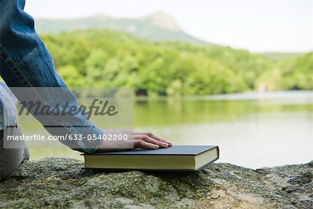 Woman sitting on rock by lake with book, cropped