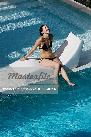 Jeune femme assise sur une chaise longue près de la piscine, balançant les pieds dans l'eau
