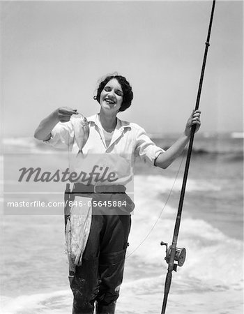 1920ER – 1930ER JAHRE LÄCHELND WOMAN STANDING IN OCEAN SURFEN TRAGEN GUMMI WATSTIEFEL HALTEN FISCH UND FLIEGE ANGELRUTE