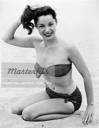 1950s YOUNG WOMAN WEARING BIKINI BATHING SUIT KNEELING ON BEACH