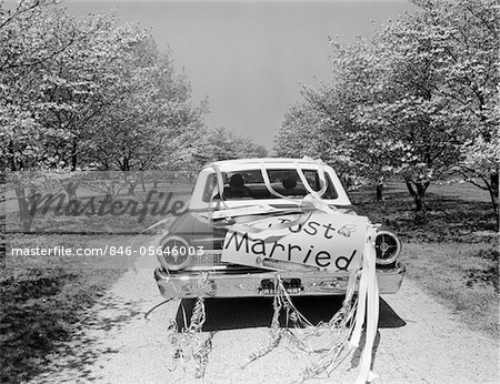 1960ER JAHRE RÜCKSEITE DES AUTO MIT JUST MARRIED SCHILDER UND STREAMER, DIE FAHRT ENTLANG DER STRAßE VON BLÜHENDEN BÄUMEN