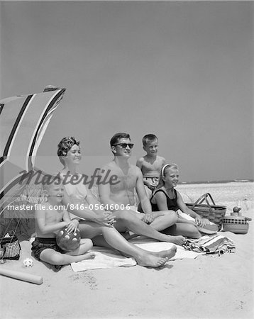 1960s FAMILY PORTRAIT MOTHER FATHER DAUGHTER AND TWO SONS SITTING ON BEACH UNDER UMBRELLA