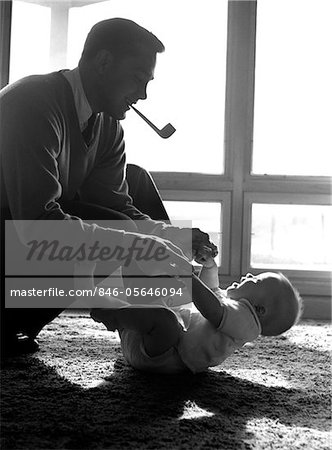 1950s FATHER PIPE IN MOUTH KNEELING DOWN TO PLAY WITH BABY LAYING ON FLOOR SUNLIGHT COMING IN THROUGH WINDOWS