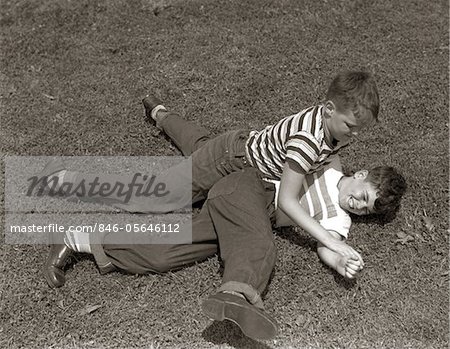 1950s TWO BOYS WEAR TEE SHIRTS BLUE JEANS PLAYING ROUGH FIGHTING WRESTLING ON THE GRASS