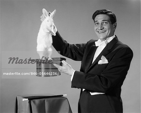1960s MAN PORTRAIT SMILING MAGICIAN PULLING WHITE RABBIT OUT OF TOP HAT
