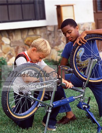 1980s AFRICAN AMERICAN AND CAUCASIAN BOYS FRIENDS WORKING TOGETHER FIXING A BICYCLE
