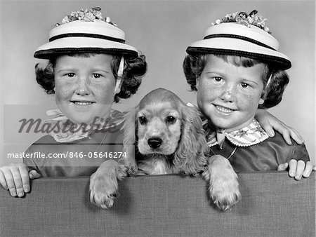 1950s - 1960s TWIN GIRLS WEARING WHITE STRAW HATS SEPARATED BY A COCKER SPANIEL PUPPY LOOKING OVER A STUDIO FENCE INSIDE