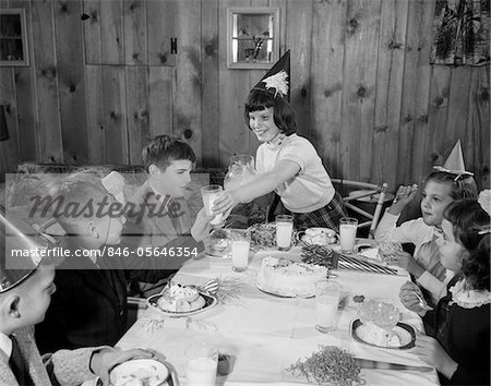 1950s GIRL POURING GLASSES MILK FOR GROUP CHILDREN AT BIRTHDAY PARTY WEARING PARTY HATS EATING CAKE