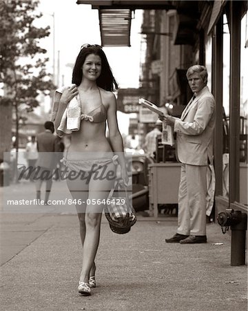 ANNÉES 1970 HOMME LORGNER SEXY SOURIANTE JEUNE FEMME MARCHANT VERS LE BAS VILLE RUE PORTANT SEULEMENT UN BIKINI MAILLOT DE BAIN