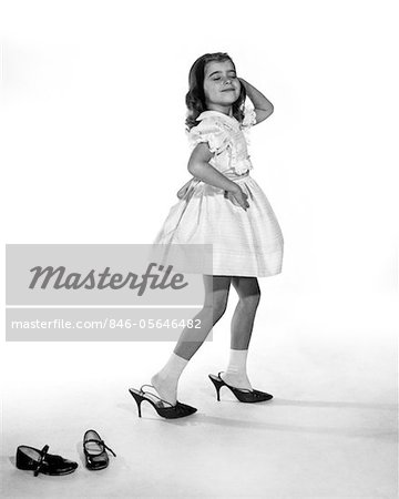 1960s GIRL MAKING GLAMOUR POSE HAVING STEPPED OUT OF HER SHOES INTO HER MOTHERS ADULT HIGH HEELS