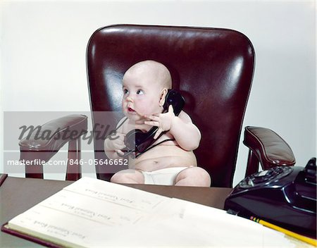 1960s CHUBBY BABY SITTING IN LEATHER CHAIR AT OFFICE DESK HOLDING TELEPHONE