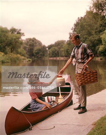 1960s YOUNG COUPLE LOADING PICNIC BASKET THERMOS INTO CANOE