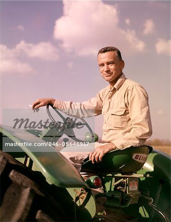 ANNÉES 1960 PORTRAIT SOURIANT HOMME AGRICULTEUR ASSIS SUR TRACTEUR VERT FUMER CIGARETTE