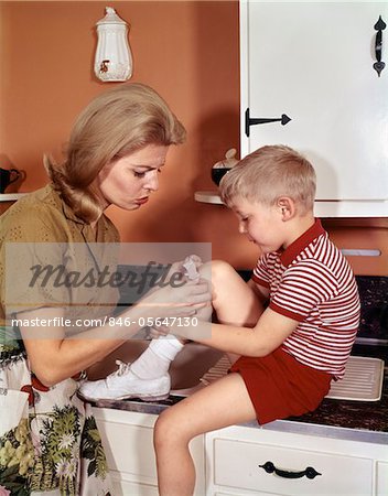 ANNÉES 1970 MÈRE DONNANT LES PREMIERS-SOINS METTANT BAND-AID ON GENOU DE FILS EN CUISINE