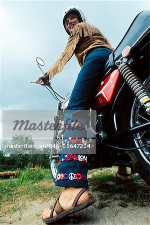 SECHZIGER JAHRE - SIEBZIGER JAHRE WOMAN ON MOTORRAD TRÄGT SANDALEN STREIFENPROJEKTION JACKE BLAU JEAN BELLBOTTOM PANTS MIT SYMBOL FÜR LIEBE UND FRIEDEN