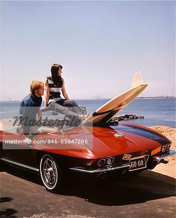 1960s TEENAGE COUPLE MAN WOMAN SITTING IN CORVETTE STINGRAY CAR WITH SURFBOARD PARKED AT BEACH