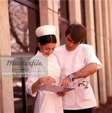 1970s DOCTOR INTERN LOOK AT CHART WITH NURSE WEARING NURSES CAP