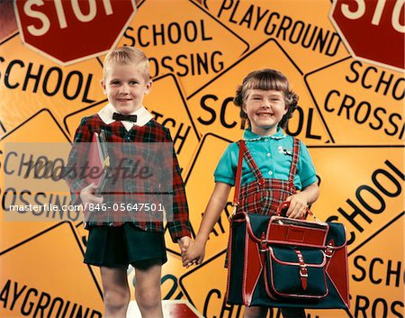 1950s GIRL AND BOY WITH BOOK BAG AND BOOKS HOLDING HANDS TOGETHER IN FRONT OF SCHOOL AND TRAFFIC WARNING SIGNS IN STUDIO
