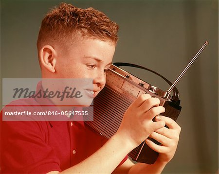 1960s SMILING YOUNG RED HAIR PRE-TEEN BOY LISTENING TO PORTABLE TRANSISTOR RADIO HELD UP TO EAR