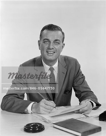 1960s SMILING HAPPY BUSINESSMAN PEN IN HAND SIGNING PAPERS AT DESK