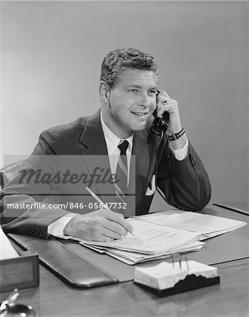 DES ANNÉES 1960, HOMME D'AFFAIRES ASSIS AU BUREAU PARLAIT AU TÉLÉPHONE SOURIANT ÉCRIT AVEC UN CRAYON