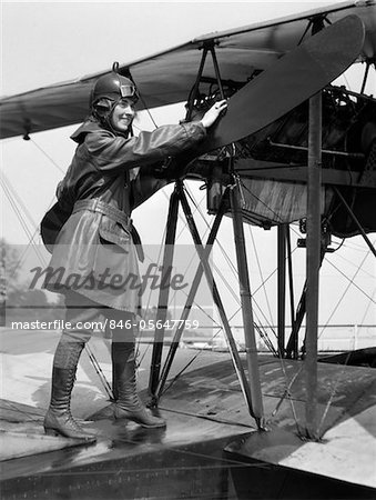 1920s SMILING WOMAN AVIATOR TURNING FLOAT BIPLANE PROPELLER WEARING LEATHER FLYING CLOTHES GOGGLES AND HELMET