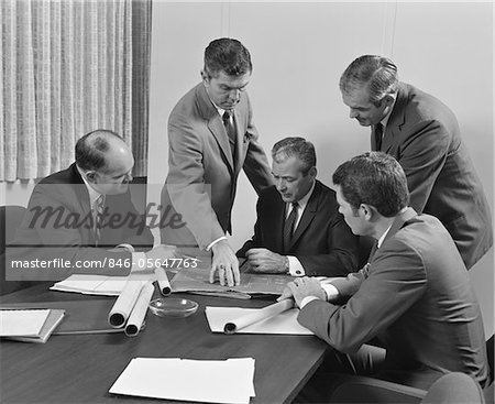 1960s 5 EXECUTIVE BUSINESSMEN AT CONFERENCE TABLE MEETING LOOKING OVER PAPERS & BLUEPRINTS