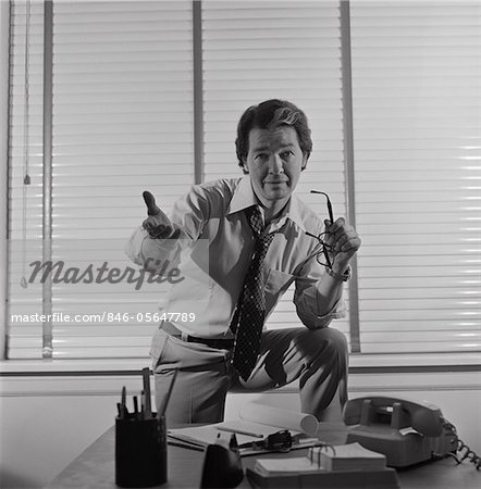 1970s BUSINESSMAN STANDING BEHIND DESK ARM RAISED IN PLEADING GESTURE