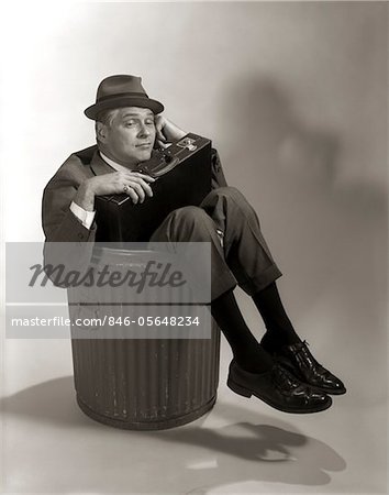 1960s SYMBOLIC BUSINESSMAN IN SUIT AND HAT HOLDING BRIEFCASE DUMPED FIRED MADE REDUNDANT LAID OFF UNEMPLOYED SITTING IN TRASH CAN