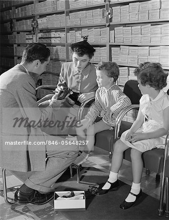 MAGASIN DE CHAUSSURES DES ANNÉES 1950 FAMILLE MÈRE DE DEUX ENFANTS EN ESSAYANT DE NOUVELLES CHAUSSURES, AIDÉS PAR LE VENDEUR