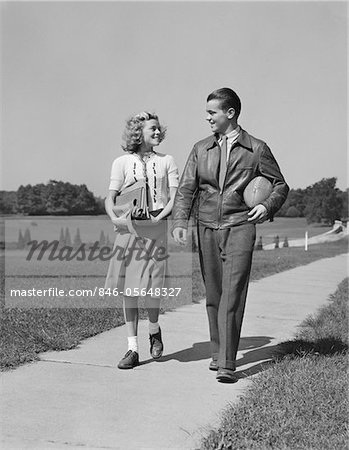 1930s - 1940s TEEN COUPLE WALKING DOWN SIDEWALK GIRL WITH SCHOOL BOOKS BOY HOLDING FOOTBALL