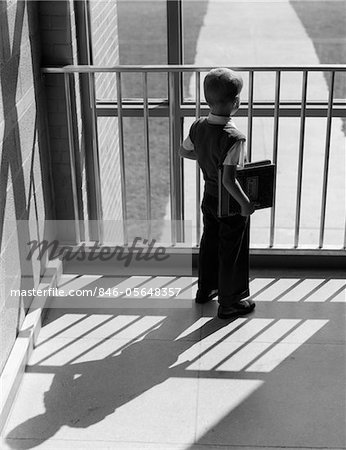 1950s SCHOOLBOY WITH BOOKS UNDER ARM LOOKING OUT WINDOW BETWEEN BARS OF RAILING SHADOW CAST BEHIND HIM