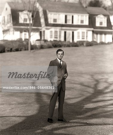 1950s MAN UPSCALE BUSINESSMAN EXECUTIVE IN GRAY FLANNEL SUIT AND TIE STANDING ON SUBURBAN HOME LAWN