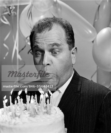 1960s MAN BLOWING OUT CANDLES ON BIRTHDAY CAKE WITH BULGING EYES & BALLOONS IN BACKGROUND