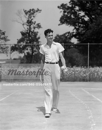 1930s MAN WALKING ACROSS TENNIS COURT HOLDING TENNIS RACKET & BALLS