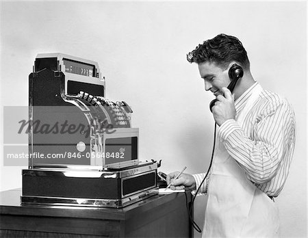 ANNÉES 1930 - ANNÉES 1940 HOMME EN TABLIER À CÔTÉ DE LA GRANDE CAISSE ENREGISTREUSE TALKING ON TÉLÉPHONE PRENANT NOTES SOURIANT
