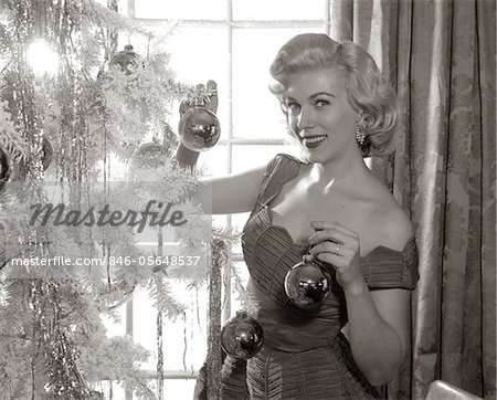 1950s WOMAN IN PARTY DRESS DECORATING INDOOR CHRISTMAS TREE HANGING BALLS ON BRANCH SMILING