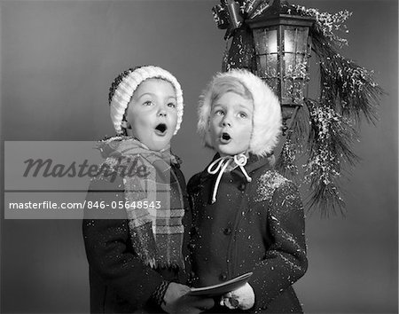 1960s BOY AND GIRL SINGING CHRISTMAS CAROL TOGETHER UNDER SNOWY OUTDOOR PORCH LIGHT