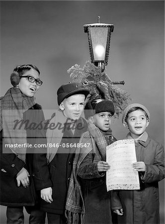 1960s FOUR MULTI ETHNIC BOYS GROUP SINGING CHRISTMAS CAROL TOGETHER STANDING BY OUTDOOR PORCH LIGHT