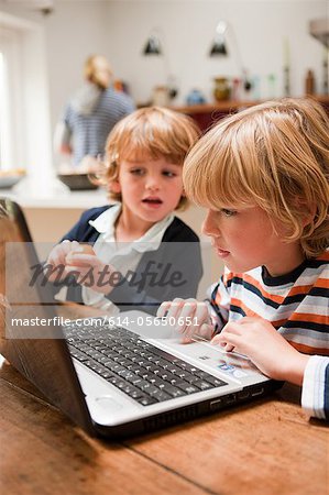 Young boy using laptop with his younger brother looking across at the monitor