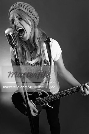 Black and white photo of a blond female screaming into an old microphone and playing electric guitar.  High contrast with film grain added.