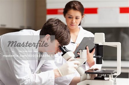 Scientist looking in a microscope while his colleague is writing in a laboratory