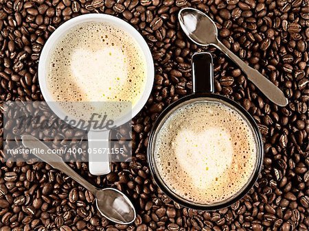 Two cups with Cafee Latte with heart shaped rosetta surrounded by coffee beans and spoons