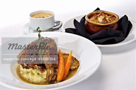 Rotisserie roasted chicken leg served over mashed potatoes with baby carrots and a gravy sauce.  A fresh green sprig of rosemary tops this off.  Warm bowls of soup are featured in the background.