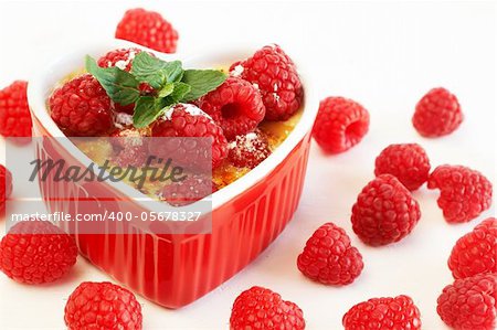 French creme brulee dessert with raspberries and mint covered with caramelized sugar in red heart shaped ramekin on white background