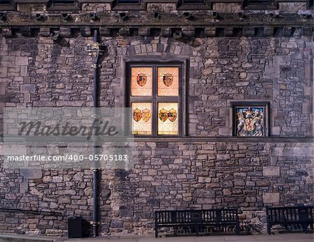 The exterior wall of the Great Hall of Edinburgh Castle at night. The stained glass windows with the emblems of past kings of Scotland are glowing in the dark next to the symbol of royalty.