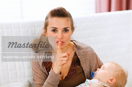 Young mama holding sleeping baby and showing shh gesture at home