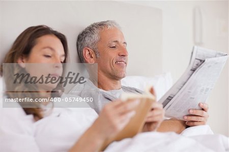 Happy woman reading a book while her husband is reading a newspaper in their bedroom