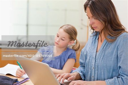 Little girl doing her homework while her mother is using notebook in a kitchen