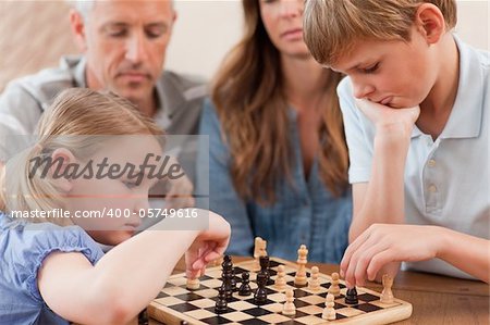 Focused siblings playing chess in front of their parents in a living room